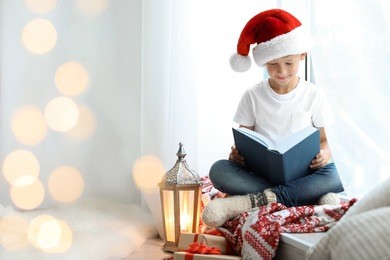 cute little child in santa hat reading christmas story while sitting on windowsill at home