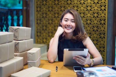 happy asian women working tablet from home on desk with postal parcel, female buyer touch screen of tablet buy presents, among gift boxes and packages. online shopping. selling online ideas concept.