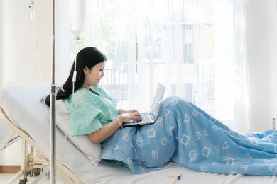 asian woman sick patient lying in a hospital bed using a laptop for relax when she recovering for ill in hospital.