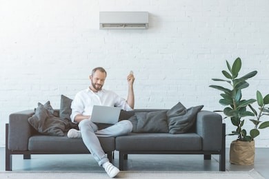 man turning on air conditioner with remote control while using laptop on sofa