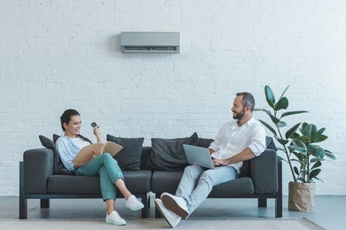 couple turning on air conditioner during the summer heat while sitting on sofa with book and laptop 