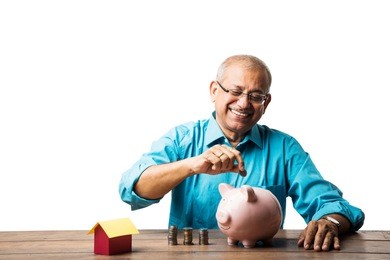 senior indian/asian man and savings concept - with piggy bank, currency notes, 3d house model, money fan, pile of coins. sitting at table