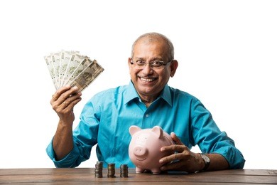 senior indian/asian man and savings concept - with piggy bank, currency notes, 3d house model, money fan, pile of coins. sitting at table