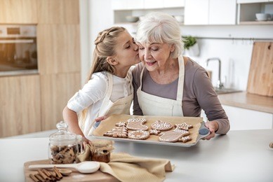 thank you, granny. cute girl is kissing cheek of her grandmother with love. senior woman is holding pray with self-baked cookies and smiling