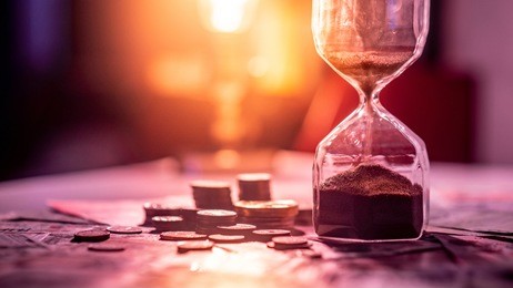 sand running through the shape of hourglass on table with banknotes and coins of international currency. time investment and retirement saving. urgency countdown timer for business deadline concept