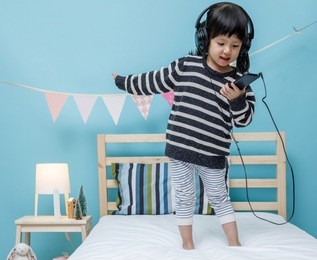 cute little girl sing a song with smartphone in her bedroom, happy asian child little girl listening the music with headphone on the bed