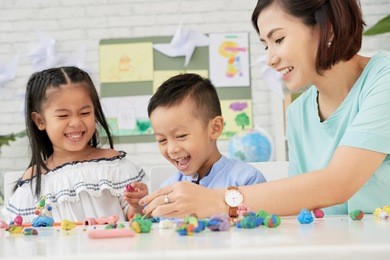 cheerful asian teacher playing with boy and girl with plasticine figurines in art class