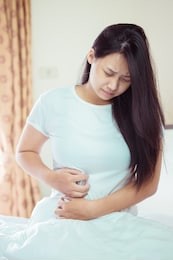 asian young woman suffering from abdominal pain while sitting on bed at home
