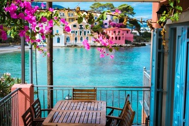 lilac fuchsia blossom flower over hotel veranda in front of turquoise colored bay of mediterranean sea and beautiful colorful houses of assos village in kefalonia, greece