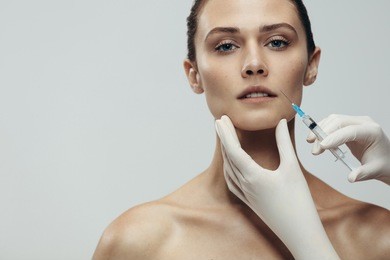 portrait of young woman getting cosmetic injection. close up of beautiful woman gets injection in her face against grey background with copy space.