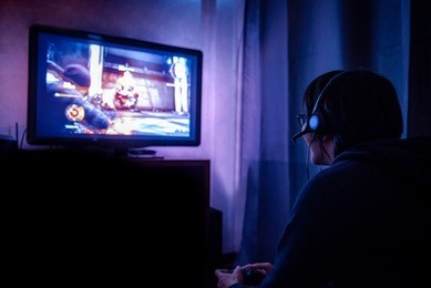 young male gamer with glasses and headset playing video game at home in the dark room using game console controller watching at led tv. gaming and entertainment concepts