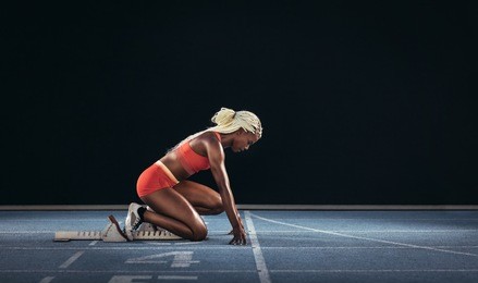 female athlete taking position on her marks to start off the run. side view of female runner  getting ready at the start line on running track on a black background.