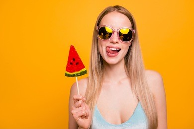 attractive nice cute childish straight-haired beautiful blonde girl, wearing blue swimsuit, color sun glasses, fooling, showing tongue out, holding candy on stick. isolated over yellow background