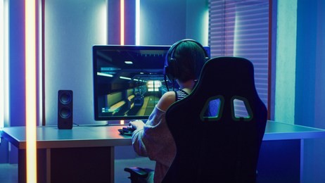 beautiful professional gamer girl playing in first-person shooter online video game on personal computer. casual cute geek girl wearing headset. dark room suddenly lit by neon lights in retro style