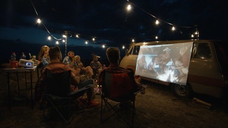 friends gathering in campsite around bonfire and watching movie with projector on van side in dark evening