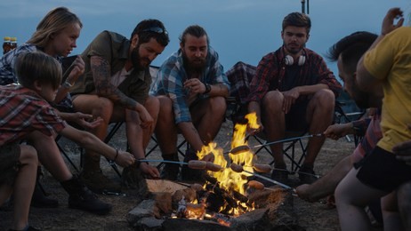 adult diverse people with childrens gathering around burning bonfire in campsite and frying sausages on skewers in flame sitting under garlands in twilight