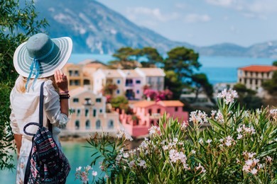 blond beautiful woman in blue sun hat and white clothes enjoying view of colorful tranquil village assos houses on sunny day. stylish female visiting kefalonia in summer time on greece travel vacation
