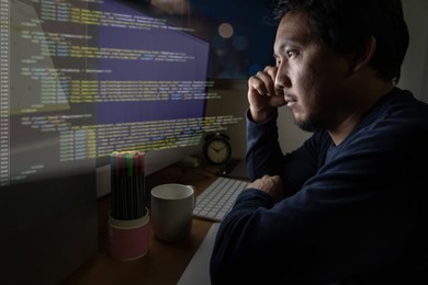 portrait of asia businessman sitting and working hard on the table with programming source code in hologram technology over front of computer desktop in work place at late with serious action