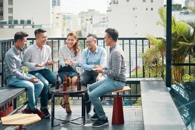 cheerful asian friends sitting on terrace with beer bottles on table and relaxing in sunlight on urban background