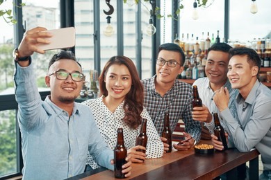 group of handsome men and charming woman with beer bottles taking selfie at table in bar smiling happily