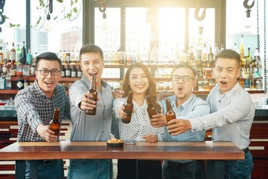 group of asian happy and bright men and woman standing at table in bar outstretching hands with beer bottles and screaming at camera