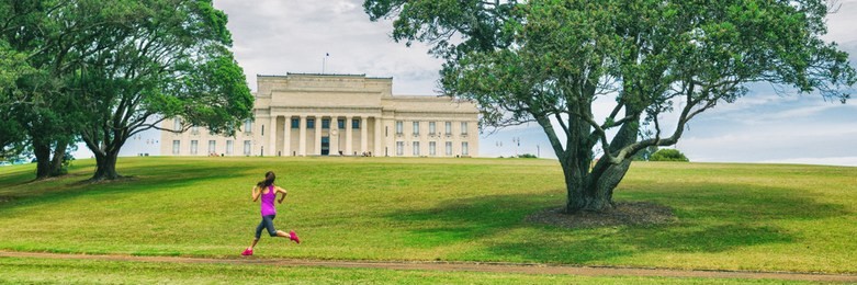 park runner woman running in auckland city banner. panoramic crop of jogging girl on green grass and trees at auckland domain park with memorial museum background. urban lifestyle.