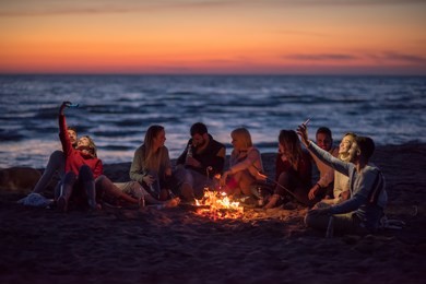 a group of friends using cell phones around the bonfire during autumn beach party drinking beer and having fun