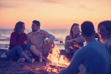 group of young friends sitting by the fire at autumn beach, grilling sausages and drinking beer, talking and having fun