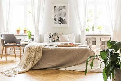 open book on a gray, wooden armchair by a cozy bed with breakfast tray in a stylish bedroom interior with natural light coming through big windows