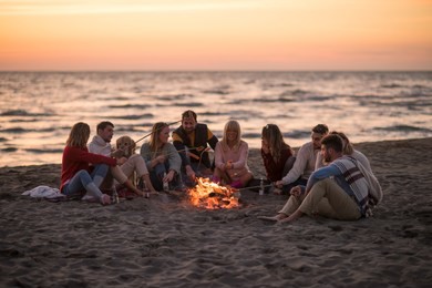 group of young friends sitting by the fire at autumn beach, grilling sausages and drinking beer, talking and having fun