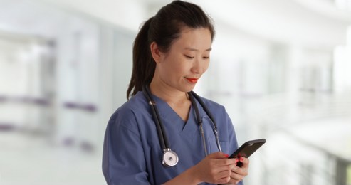 happy young asian doctor in scrubs texting with smartphone inside empty hospital