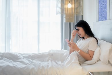 asian woman in bed using mobile phone and feeling eye pain at bedroom in home.