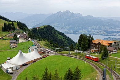 a sightseeing train traveling on the cogwheel railway through green grassy meadows on mt. rigi, with rugged pilatus peaks among majestic mountains in background on a cloudy summer day in switzerland