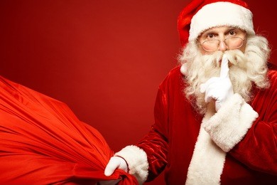 portrait of santa claus with huge red sack keeping forefinger by his mouth and looking at camera