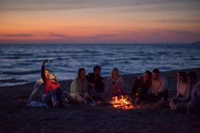 a group of friends using cell phones around the bonfire during autumn beach party drinking beer and having fun