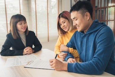 happy young asian couple and realtor agent. cheerful young man signing some documents while sitting at desk together with his wife. buying new house real estate. signing good condition contract.