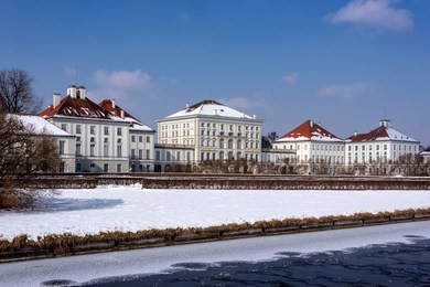 germany, bavaria, munich, nymphenburg palace: panorama view of winter scene with main building entrance of the famous castle, snow, ice, canal, blue sky in the bavarian capital.