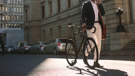 cropped image of businessman walking on city street with a bicycle and talking on mobile phone. man in formal wear going to work with cycle using phone.