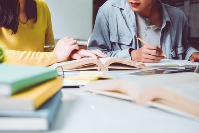 high school or college using laptop while sitting at table. group students studying and reading with books in library. students helps friend catching up and learning tutoring. education concepts
