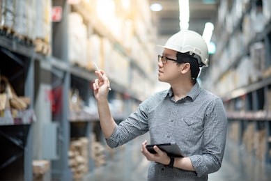 young asian man worker wearing safety helmet and eyeglasses doing stocktaking of product in cardboard box on shelves in warehouse by using digital tablet and pen. physical inventory count concept