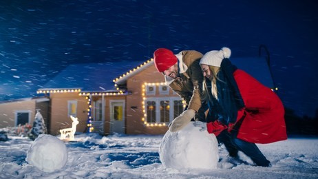 happy young couple makes a snowman, both rolling big snowball. family having fun on one winter evening. in the background house decorated with garlands.