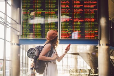 theme travel and tranosport. beautiful young caucasian woman in dress and backpack standing inside train station or terminal looking at a schedule holding a red phone, uses communication technology.