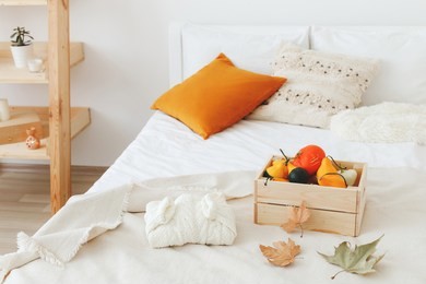 home decor. cozy fall bedroom interior: white wall, wooden rack, bed with white linen, light beige plaid, orange pillows,  wooden box with pumpkins, dry autumn leaves, sweater. autumn decoration.
