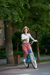 smiling pretty girl in red skirt holding flowers in her hands is riding blue bicycle down paved city street surrounded with green trees. beautiful positive female peonies stroll turquoise bike summer