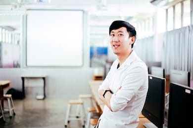 portrait of young handsome smiling asian male teacher in computer lab classroom background