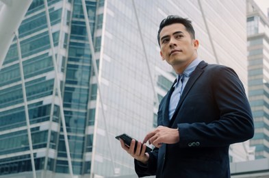 young handsome asian business man wearing modern black suit reading information about finance news with mobile smart phone in building city background, network technology, internet, successful concept