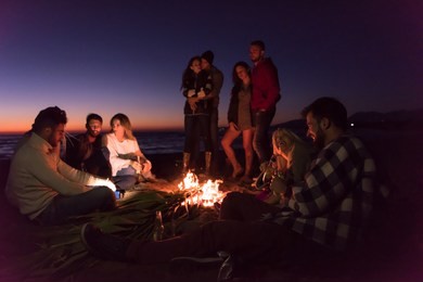 happy carefree young friends having fun and drinking beer by bonefire on the beach as the sun begins to set