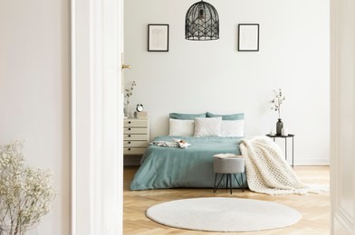 a view through an open door into a sunny bedroom interior with sage color linen and cushions on a bed, blanket, drawer cabinet, soft stool and a round rug. real photo