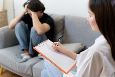 female professional psychologist is conducting a consultation to stressful male patient sitting on sofa in her office. she is writing down on a paper. psychological consultant concept.