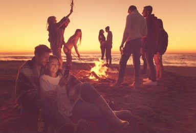 couple using cell phone during autumn beach party with friends drinking beer and having fun